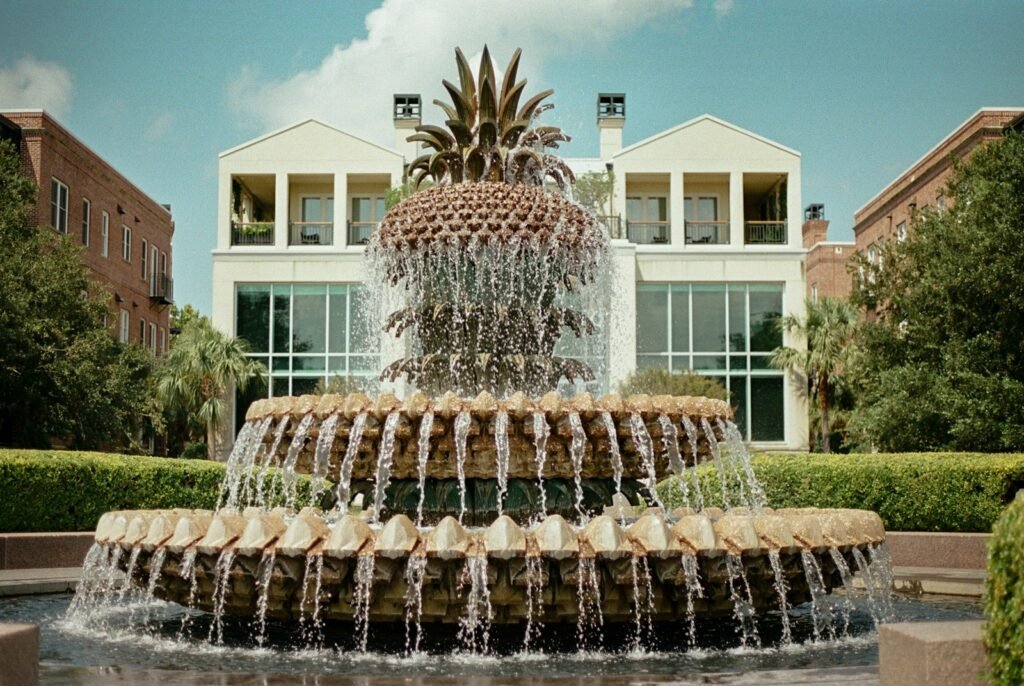 The pineapple-shaped Pineapple Fountain at Waterfront Park in Charleston with water falling over its tiers and white buildings behind