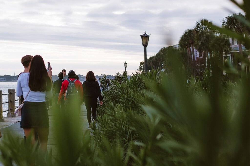 People strolling along a waterfront boardwalk lined with palmetto plants and a lamp post at dusk in Charleston