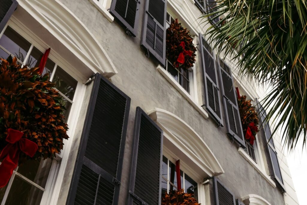 Magnolia-leaf wreaths with red velvet bows hanging in the windows of a historic Charleston house beside a palmetto frond