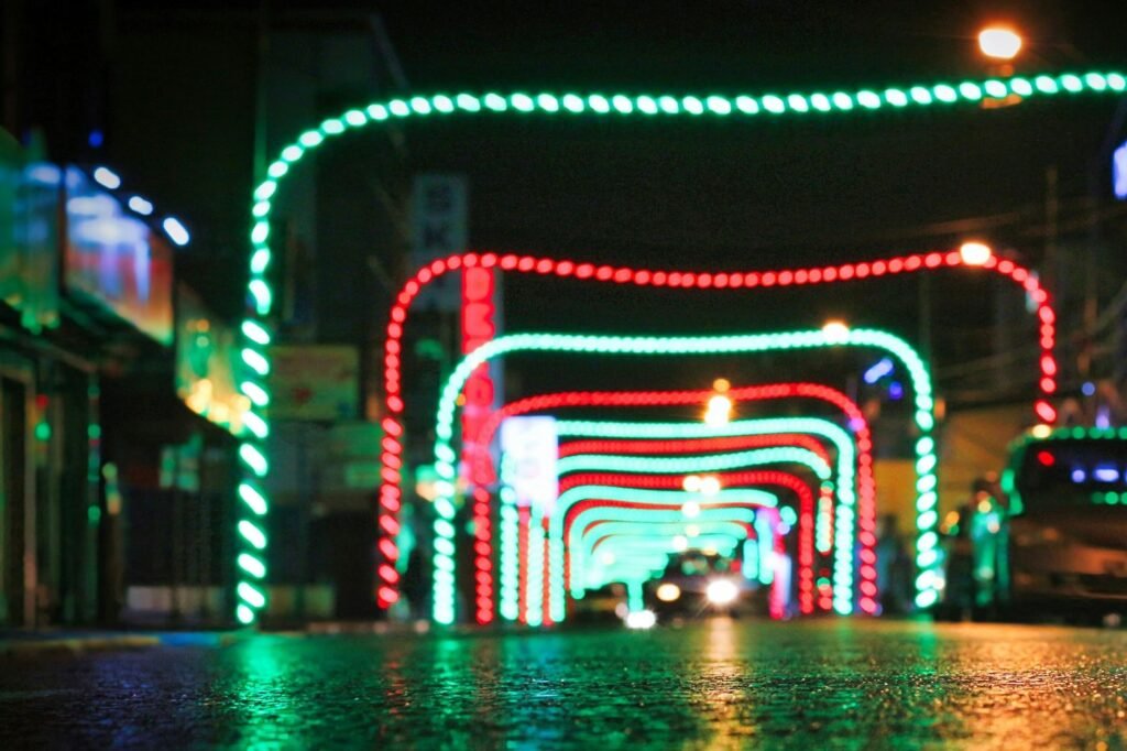 A car driving toward a tunnel of red and green Christmas light arches over a wet road at night