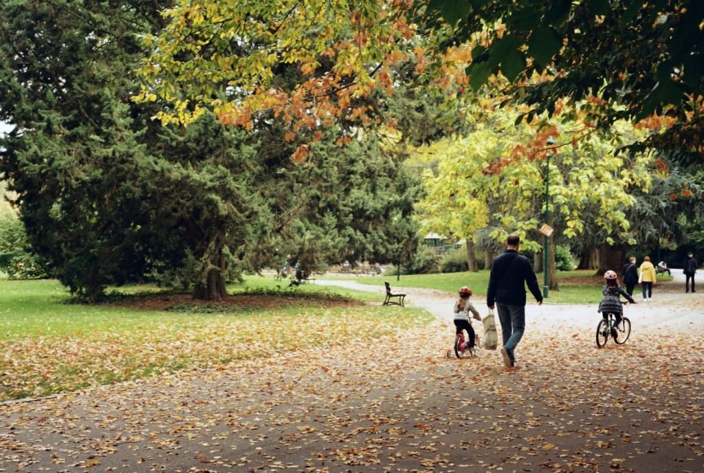 A man walks along a leaf-covered park path while two children ride small bikes beneath autumn trees