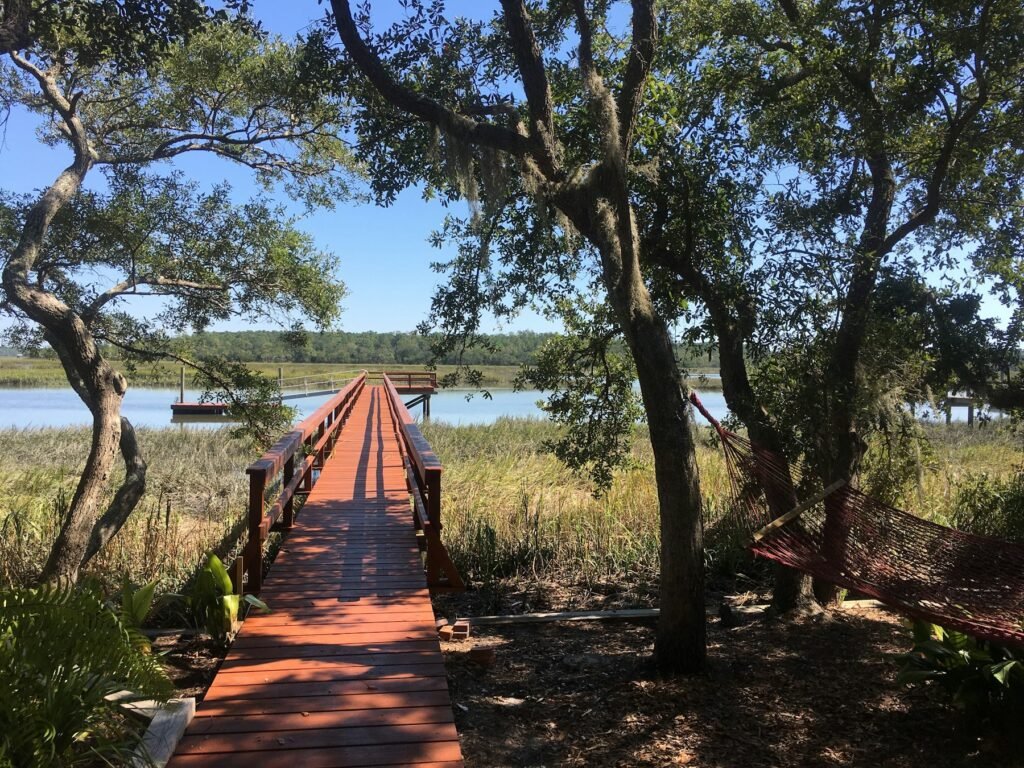 Wooden walkway leading to a dock over a tidal creek, framed by live oaks and marsh grass