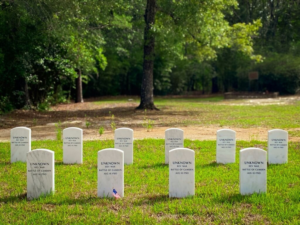 Rows of white military-style grave markers on a green lawn, with a small flag and trees behind