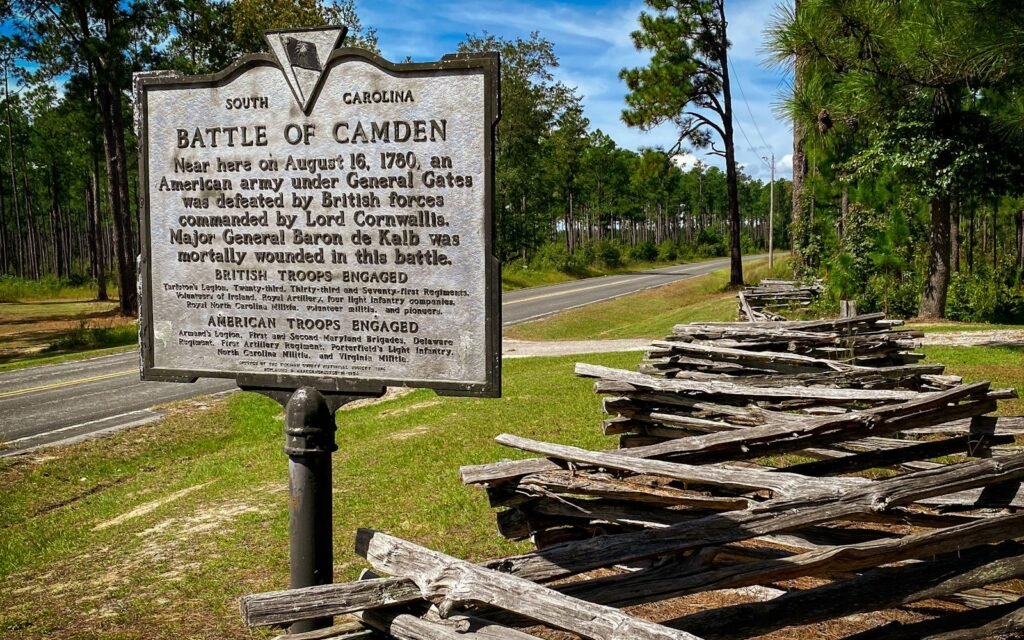 Battle of Camden historical marker beside a split-rail fence and pine woods on a sunny day
