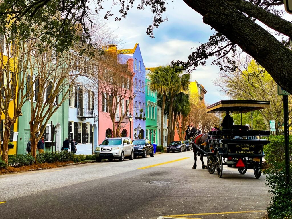 Pastel-colored row houses with palm trees on a Charleston street, a horse-drawn carriage passing at right