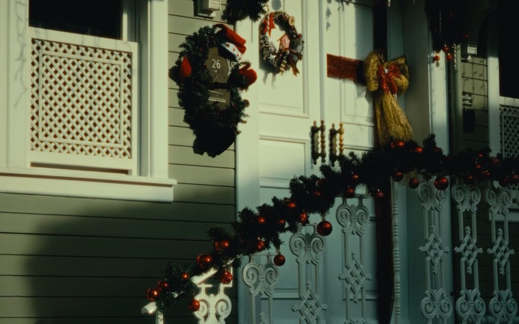 Christmas wreaths and a garland with red ornaments on the white trimmed porch of an old wooden house