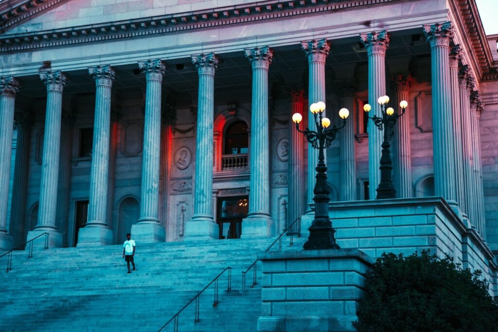 Tall fluted columns and stone steps at dusk on the front of the South Carolina State House with ornate lamp posts and one person on the steps