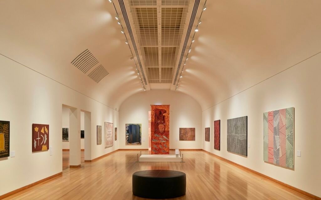 Long bright gallery with framed paintings on white walls and a round bench in the middle
