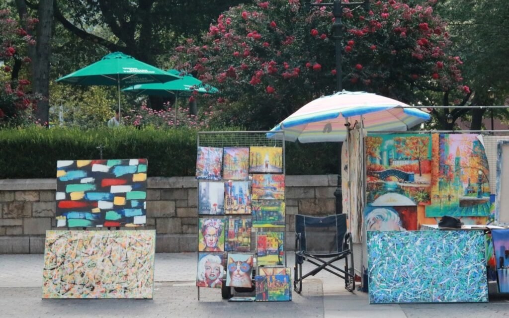 Outdoor art stalls with colorful paintings on easels under umbrellas beside a stone wall
