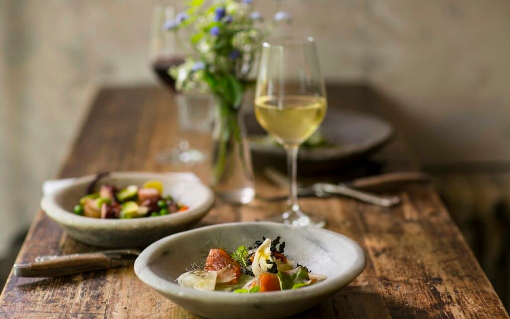 Glass of white wine, a small vase of flowers and two plated dishes on a rustic wooden table