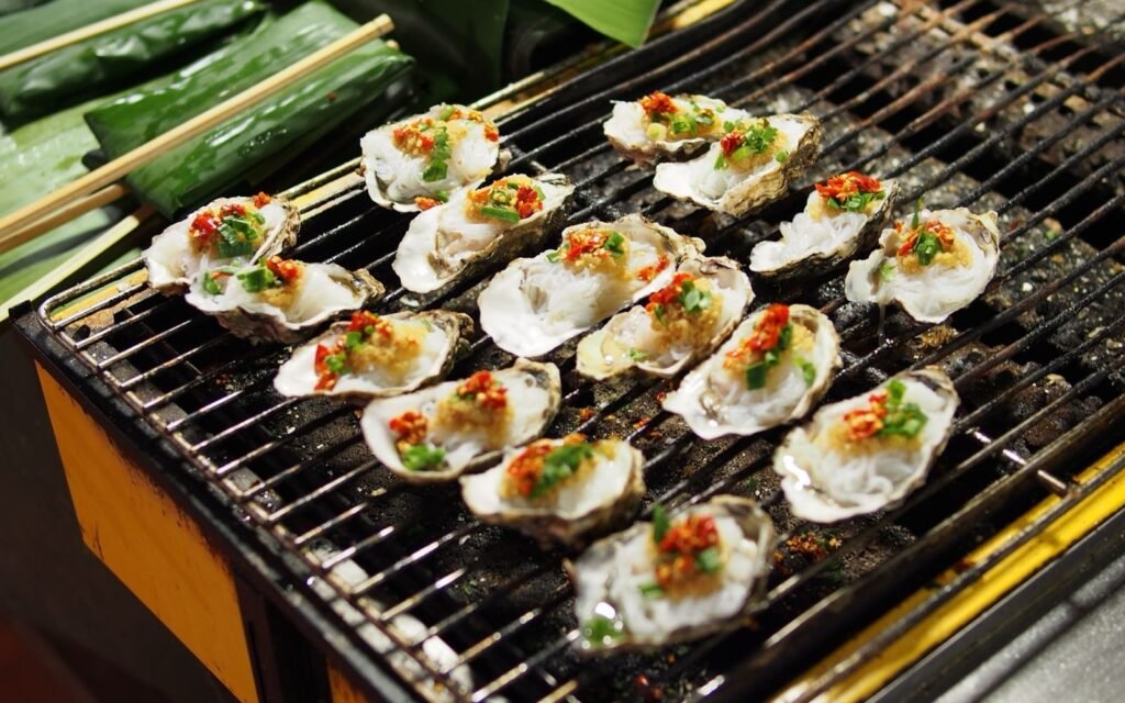 Open oysters topped with chopped peppers cooking on a metal grill rack