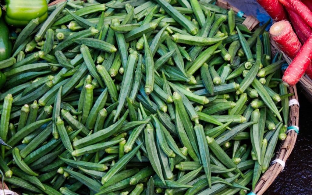 Basket heaped with fresh green okra pods beside red carrots at a vegetable market