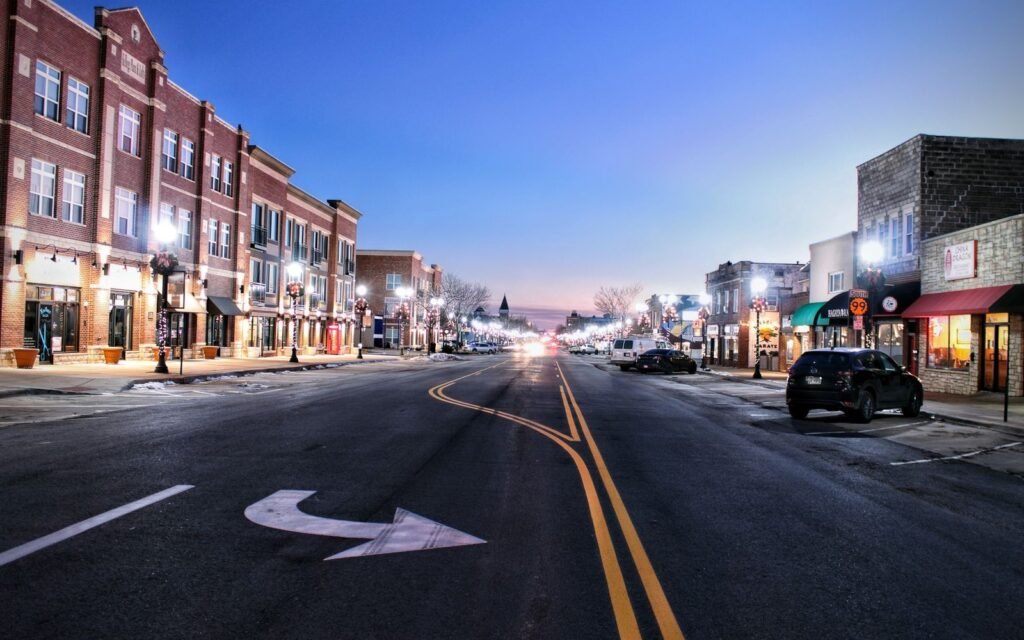 Small-town main street of brick storefronts and lit street lamps at twilight with an empty road