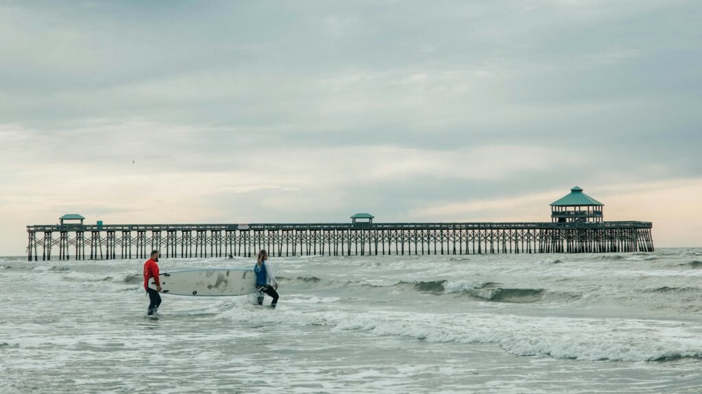 Long wooden fishing pier at Folly Beach seen from the surf, two surfers standing in the waves under a gray sky