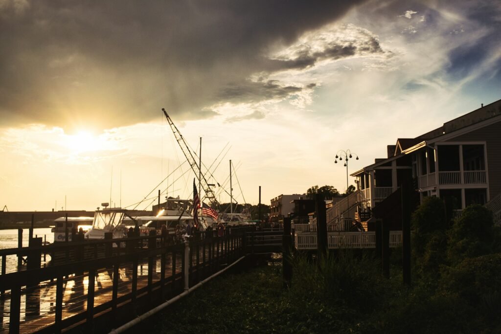 Waterfront boardwalk at sunset with stilted riverside houses and boat masts silhouetted against dark clouds