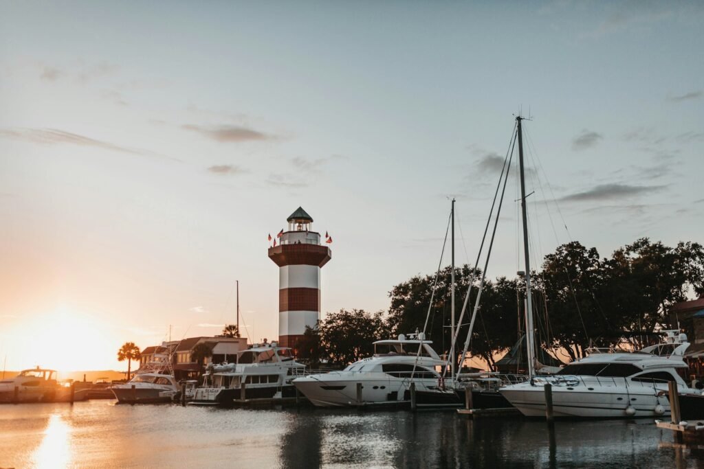 Red-and-white striped lighthouse among docked yachts and sailboats at a harbor at sunset with live oaks behind