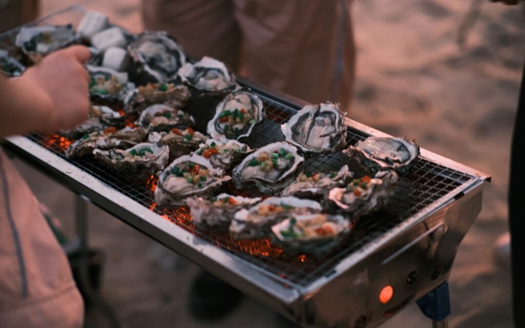 Oysters in the shell roasting over glowing coals on a portable outdoor grill at dusk
