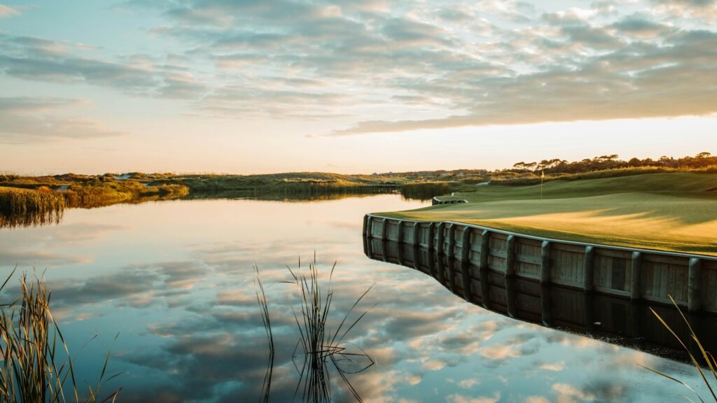 Golf green with a timber retaining wall beside a reflective marsh lake at sunset, clubhouse in the distance