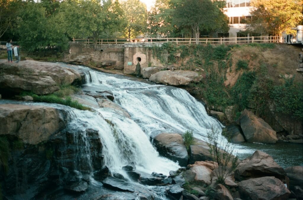 Waterfall cascading over broad rocks into the Reedy River beneath trees with city buildings above