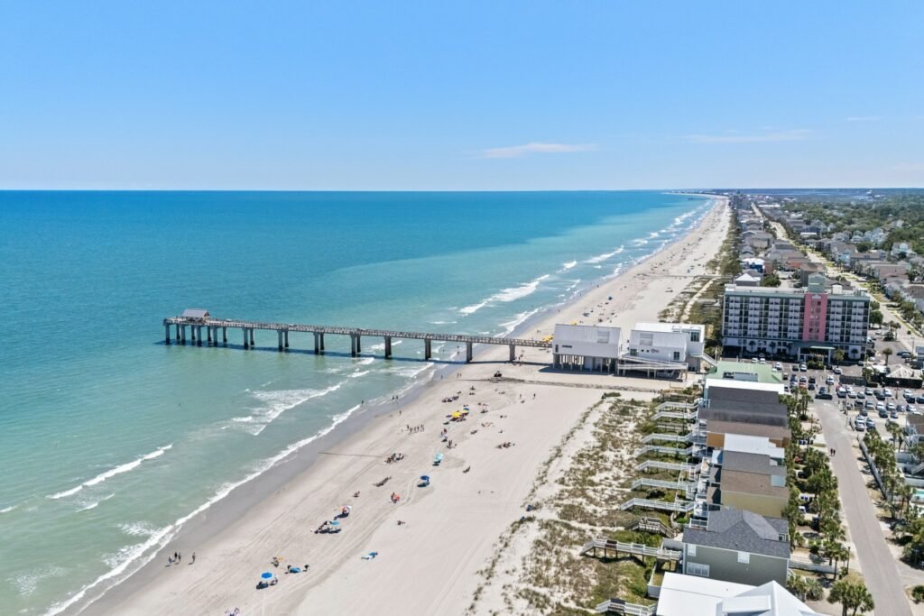 Aerial view of an oceanfront beach with a fishing pier extending into the surf and beachfront buildings