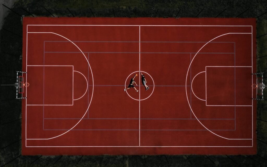 Overhead view of an outdoor red basketball court with white lines and two tiny players