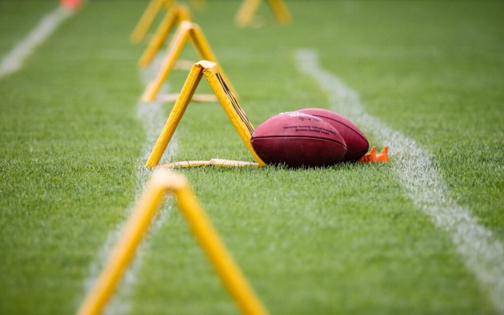 Two footballs lying on green turf beside yellow agility hurdles and a white sideline