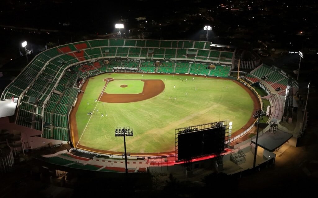 Aerial view of a lit baseball stadium at night with green seats and a dark scoreboard