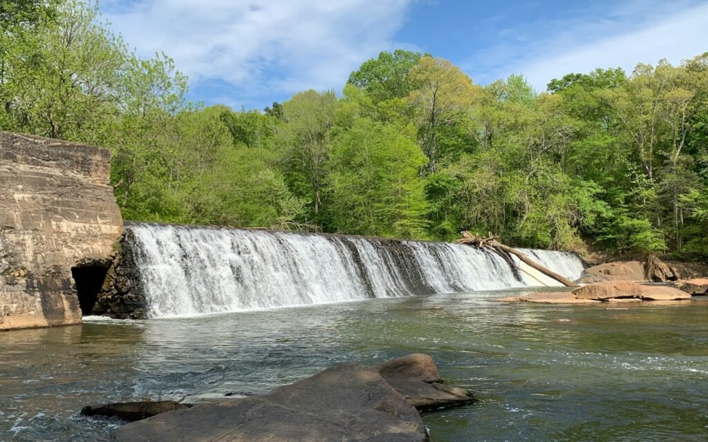 Wide waterfall spilling over a rock ledge beside an old stone wall into a river with green trees