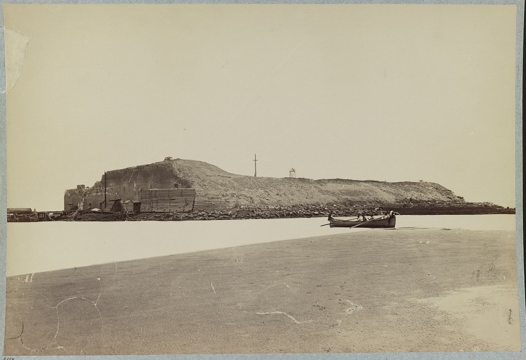 Battered walls of Fort Sumter seen across the water from a sandbar, with a rowboat in front