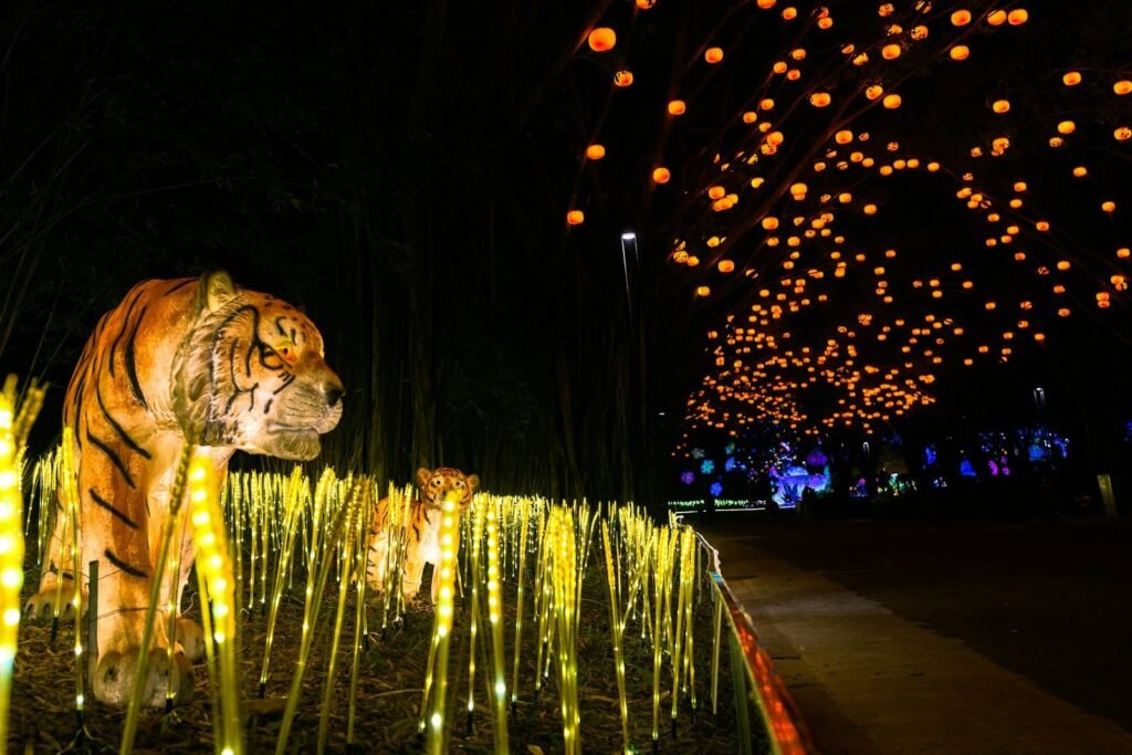 Glowing tiger lantern figures among lit wheat-shaped lights, with hundreds of orange lanterns hanging over a path at night